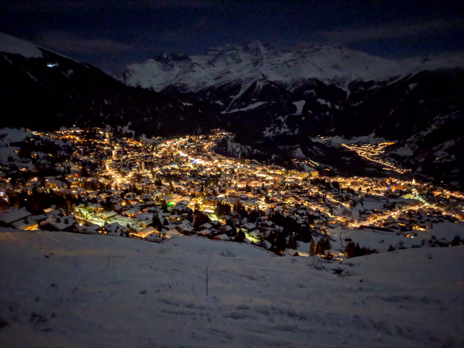 Verbier village lit up at night, seen from the slopes above, with the peaks of the 4 Vallees behind