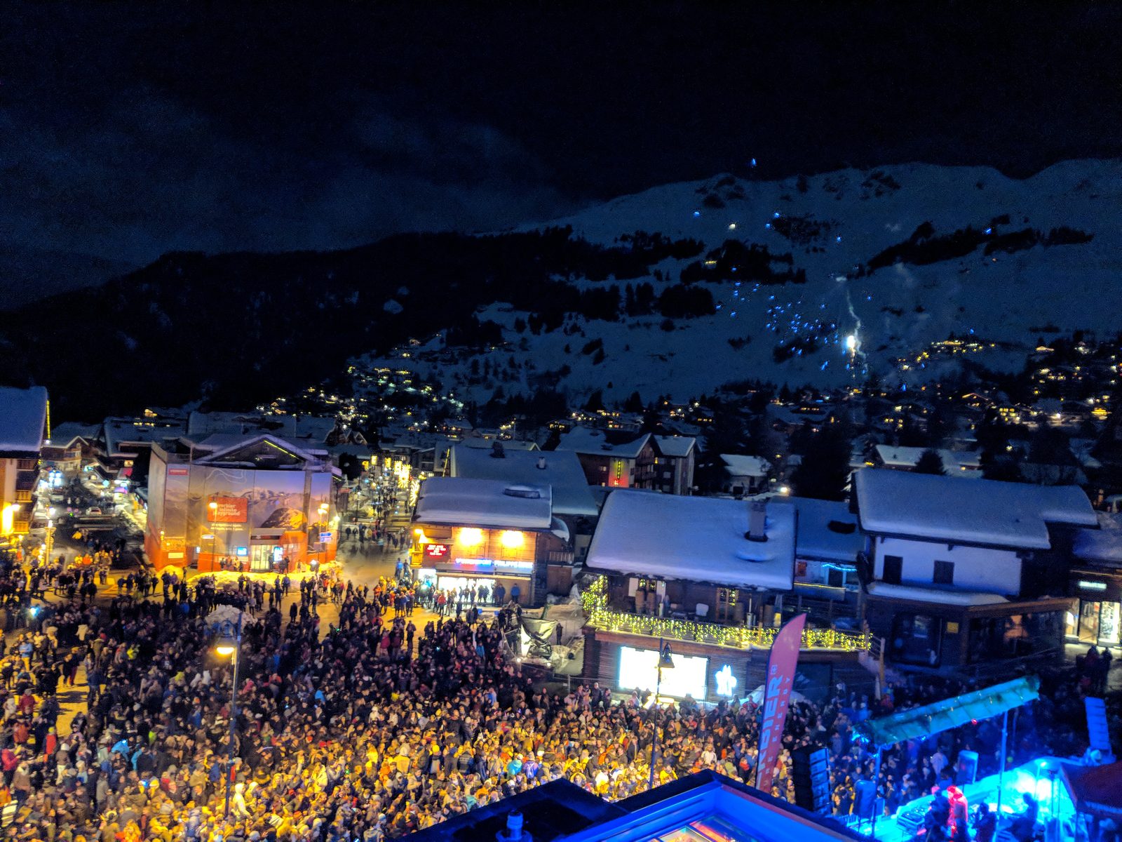 New Year's Eve crowds filling Verbier's Place Centrale at night, photographed from Hotel Farinet, with fireworks smoke over the mountain behind